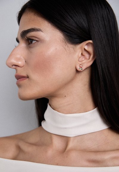 Cercei cu surub din aur, cu un design în formă de trandafir roșu și un tulpină verde. Modelul poartă o guleră de culoare deschisă și are părul lung, de culoare închisă.