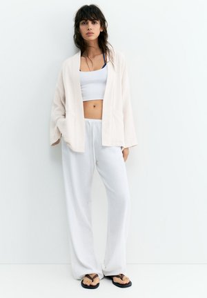 Kobieta stoi przy białej ścianie, nosząc jasnobeżowy otwarty kardigan, biały krótki top, luźne białe spodnie i czarne klapki.