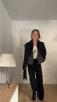 Frau lächelt und geht drinnen, trägt schwarze Hose, weißes Oberteil, grauen Blazer, schwarzen Schal und silberne Halskette, neben einer Lampe und einem Holztisch.