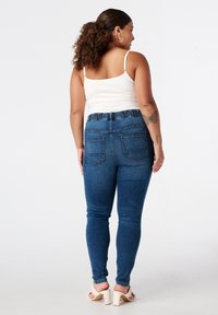 Jean skinny en denim bleu foncé, avec une taille élastique et deux poches arrière, associé à un débardeur blanc et des talons neutres.