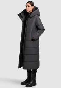 Cappotto imbottito lungo di colore grigio scuro con cappuccio, caratterizzato da una chiusura con zip, tasche laterali, texture trapuntata e taglio dritto.