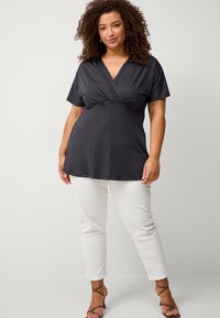 Haut noir à manches courtes avec un col en V et une taille froncée, associé à un pantalon blanc ajusté et des talons bruns à lanières, le tout sur fond neutre.