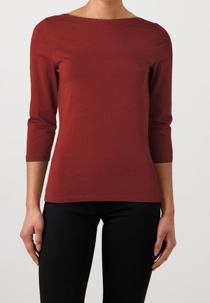 Personne portant un haut brique à manches trois-quarts avec un col bateau et un pantalon noir, debout devant un fond clair uni.