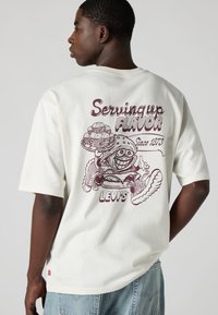 Λευκό βαμβακερό T-shirt με μεγάλο μωβ γραφικό στην πίσω πλευρά, που απεικονίζει έναν χαμογελαστό χαρακτήρα με φαγητό, μαζί με το κείμενο "Serving up Flavor".