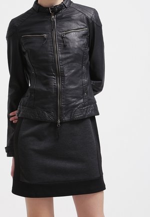 Veste en cuir noire avec fermeture éclair à l'avant et poches zippées sur la poitrine portée sur une jupe noire, mains du modèle derrière le dos, fond clair uni.