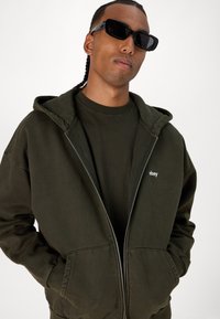 Tamnozelena zip-up hoodie od mješavine pamuka s velikim prednjim džepovima, mekom teksturom, i "obey" logom na prsima. Nosjena preko odgovarajuće majice.