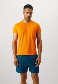 Mandig atletisk outfit med en lys orange t-shirt med korte ærmer og et hvidt logo, kombineret med dyb teal shorts. Glat stoffeeksstruktur.