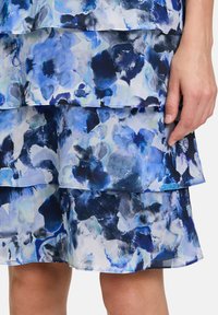 Niebieska sukienka w kwiatowy wzór z falbaniastymi warstwami, wykonana z lekkiego materiału, w odcieniach niebieskiego, granatowego i z akcentami zieleni.