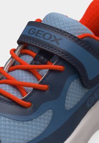 Κοντινό πλάνο μπλε και ναυτικού sneaker με έντονα πορτοκαλί κορδόνια και ναυτικό ιμάντα με την επωνυμία "GEOX" στο επάνω μέρος.