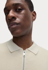 Lyse beige, tekstureret strik polo med lynlås foran og en ribbet krave. Nærbillede der viser ansigtstræk og hudstruktur.