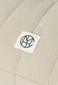 Beige teksturert pute med quiltet design. Har et hvitt stoffmerke som viser en svart sirkulær logo og initialene "MYC."
