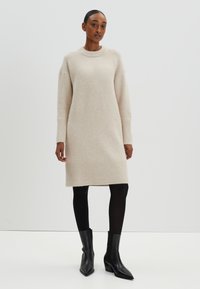 Robe-sweater beige en tricot avec des manches longues, des poignets et un bas côtelés. Portée avec des collants noirs et des bottines à bout pointu. Texture lisse, coupe décontractée.