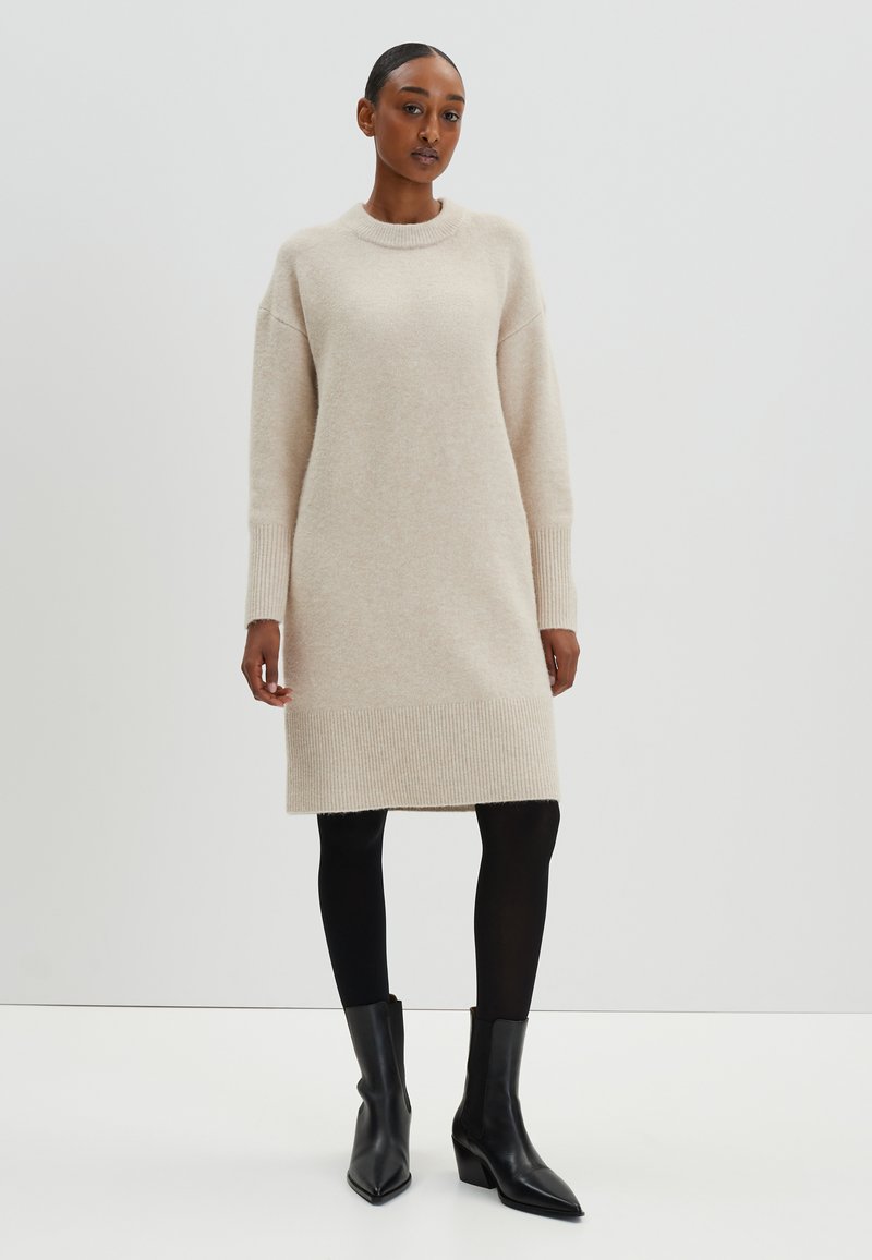 Beige gestricktes Kleid mit langen Ärmeln, gerippten Bündchen und Saum. Getragen mit schwarzen Strumpfhosen und spitzen Knöchelstiefeln. Glatte Textur, lockere Passform.