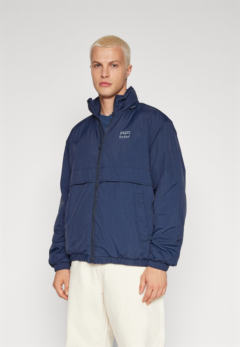 Tamsiai mėlynas lengvas švarkas su aukšta apykakle, priekiniais užtrauktukais ir elastingomis rankovėmis. Yra krūtinės kišenė su logotipo uždėklai. Lygus paviršius.