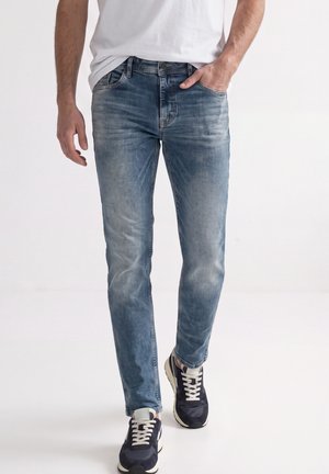 Mand iført falmede blå jeans og sorte sneakers går mod en ensfarvet hvid baggrund.