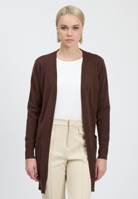 Brune lange cardigan met open voorkant, lichtgewicht materiaal, twee voorzakken, gedragen over een wit shirt en beige broek.