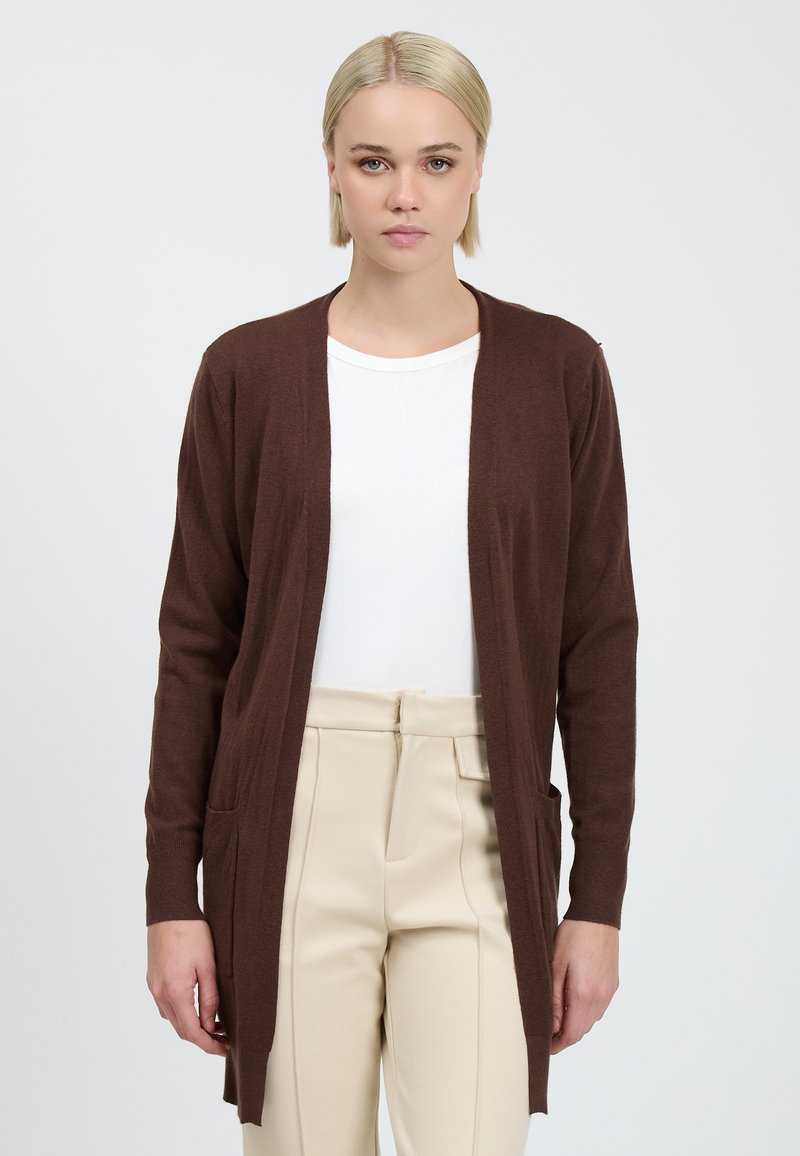 Brune lange cardigan met open voorkant, lichtgewicht materiaal, twee voorzakken, gedragen over een wit shirt en beige broek.