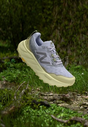 Svetlosivé a béžové bežecké topánky New Balance s hrubou podrážkou, šnurovaním a značkou „Fresh Foam“, zobrazené vo vzduchu nad trávnatým terénom.