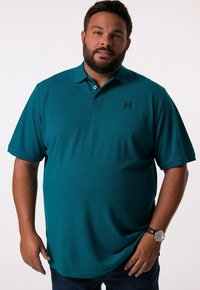 Tyrkysové polo tričko s krátkými rukávy, tříknoflíkovou légou a jemným logem na hrudi. Hladká tkanina a pohodlný střih, zkombinované s tmavými džíny.