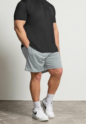 Mand iført sort kortærmet skjorte, lysegrå mesh-shorts, hvide sokker og hvide Nike-sneakers, står op ad en almindelig væg.