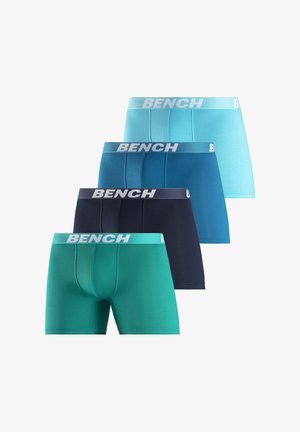 Mann trägt türkisfarbene Boxershorts mit weißem "BENCH"-Logo auf dem Bund und steht vor einem schlichten hellen Hintergrund.