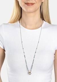 Perlesmykke med transparente og blå sten samt en gylden ring som accent, vist på en lys baggrund og båret over en hvid skjorte.