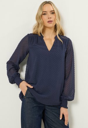 Donna che indossa una blusa blu navy trasparente a maniche lunghe con motivo a pois in rilievo e polsini arricciati, abbinata a jeans blu scuro, in piedi contro un muro bianco.