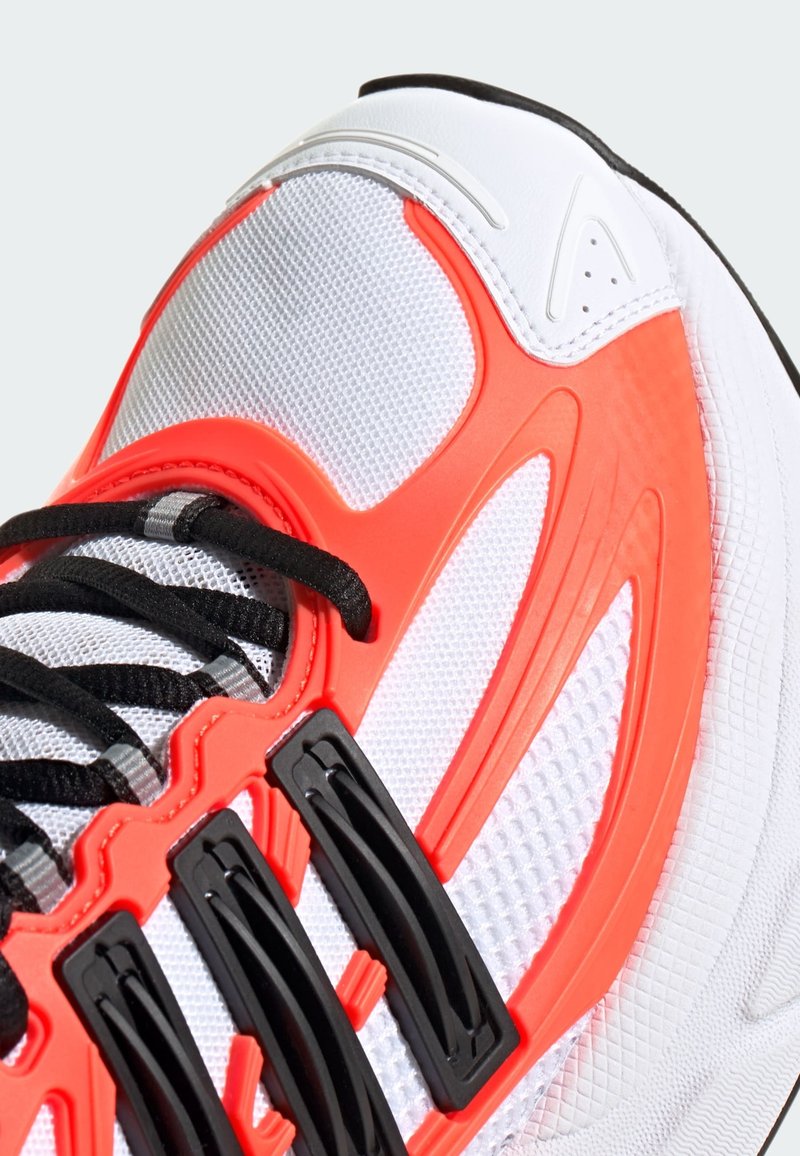 Sportsko med en hvid mesh-overdel, røde plastdetaljer, sorte snørebånd og teksturerede sorte hardwaredele. Den har et buet og strømlinet design.