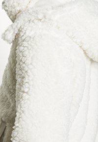 Bílá načechraná fleecová bunda s texturovaným, plyšovým povrchem a kapucí. Vlastnosti zahrnují vázané detaily poblíž výstřihu. Vhodná pro chladnější počasí.