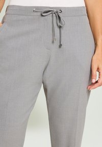 Graue Hose mit einem Kordelzugbund, verziert mit Metallakzenten und seitlichen Taschen. Glatte Textur, tailliertes Design und zulaufende Form.