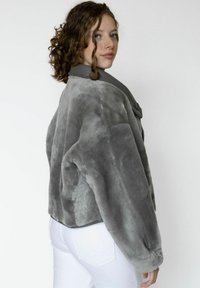 Harmaa tekoturkistakki, jossa leveä, pyöreä kaulus ja muhkeat hihat. Pehmeä pinta, napit edessä. Valkoiset housut näkyvissä alapuolella.