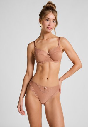 Vrouw met haar in een knot, gekleed in een bijpassende huidkleurige bh en slipje met kant, staand met één hand op de heup tegen een effen grijze achtergrond.