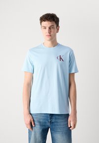 Světle modré bavlněné tričko s krátkým rukávem a kulatým výstřihem, s malým vínově červeným logem "cK" na levé části hrudníku.
