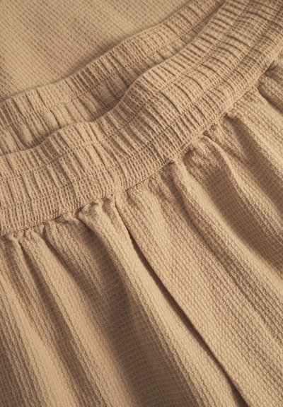 Tissu texturé beige avec un motif gaufré. Dispose d'une taille élastique avec des détails froncés et de plis doux et drapés.