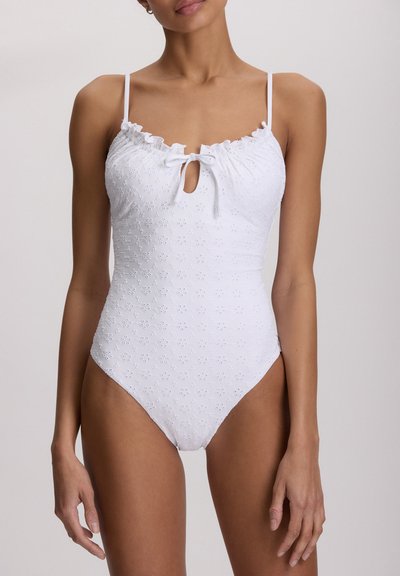 Γυναίκα που φοράει λευκό ολόσωμο μαγιό χωρίς μανίκια με φλοράλ σχέδιο σε διάτρητο ύφασμα και μικρή λεπτομέρεια δέσιμου μπροστά σε σχήμα φιόγκου.