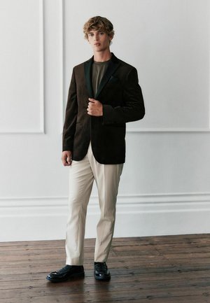 Fiatal férfi fekete blézerben, sötét ingben, bézs nadrágban és fekete alkalmi cipőben áll egy fa padlón, fehér kazettás fal előtt.