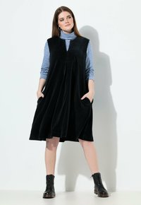 Schwarzes Cordkleid mit hohem Kragen, seitlichen Taschen und ausgestelltem Schnitt, getragen über einem blau-weiß gestreiften Rollkragenpullover. Schwarze Stiefel.