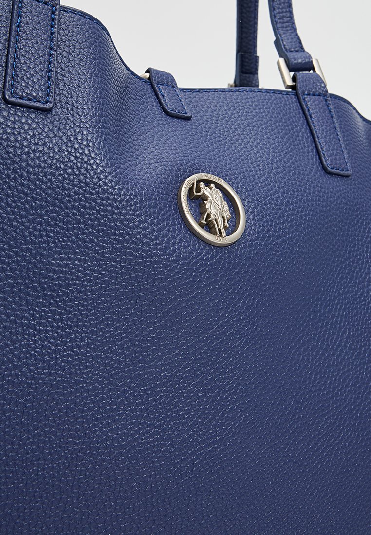 Bolso de mano de cuero texturizado azul con un prominente emblema redondo plateado que lleva una figura y asas de hombro resistentes.