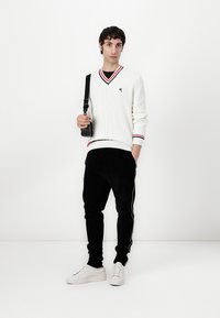 Weißer gestrickter Pullover mit V-Ausschnitt und rot/schwarzen Streifenakzenten, kombiniert mit schwarzen gerippten Jogginghosen und weißen Sneakers. Schwarze Umhängetasche.