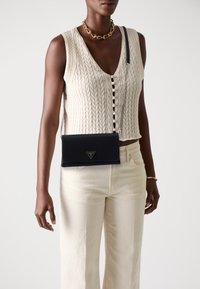 Sac en cuir noir porté en travers avec une finition texturée, arborant un logo triangulaire et une bandoulière en chaîne argentée, porté avec un haut en tricot crème.