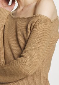 Schulterfreier, langärmeliger Pullover aus hellbraunem Strick. Mit einem gerippten Muster und einem weiten Ausschnitt, der den Schulterbereich betont.