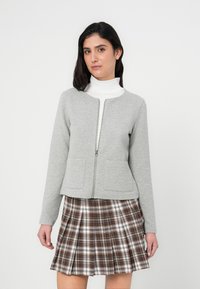 Γκρι μπουφάν με φερμουάρ και δύο μπροστινές τσέπες, σε συνδυασμό με καρό πλισέ φούστα σε καφέ αποχρώσεις με λευκές λεπτομέρειες. Ύφασμα λείο και υφή.