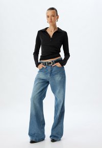 Schwarzes langärmliges Poloshirt mit Kragen, kombiniert mit hellblauen Weitbeinjeans. Abgerundet mit einem schwarzen Gürtel mit einer silbernen Schnalle.