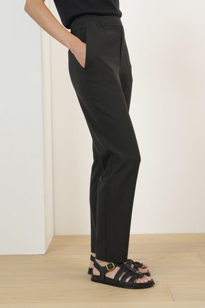 Black tapered pants with an elastic waistband and side pockets, paired with braided black sandals featuring a gold buckle accent.