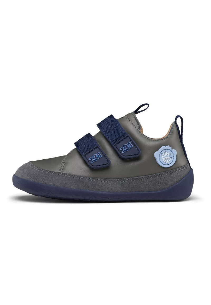 Grauer Lederschuh mit marineblauen Akzenten, zwei Klettverschlüssen und einer Gummisohle. Mit einem runden blauen Logo-Detail an der Seite.
