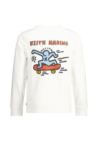 T-shirt blanc à manches longues avec un personnage de dessin animé bleu sur un skateboard, avec "KEITH HARING" imprimé au-dessus en lettres épaisses orange et noires.