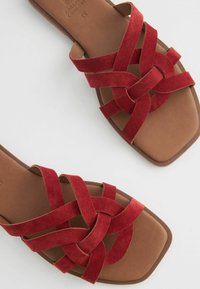 Røde ruskinds slide-sandaler med et knyttet stroppedesign. Firkantet tå og brun sål. Den glatte tekstur står i kontrast til de bløde stropper.