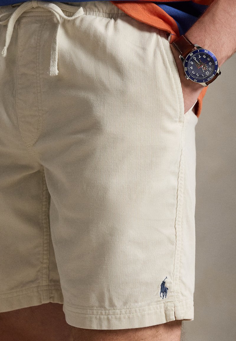 Mann trägt beige Cord-Shorts mit Kordelzug und blauer Polo-Logo, oranges Hemd und Armbanduhr mit braunem Lederarmband.