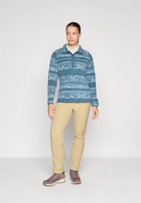 Fleece pulli i blå med geometriske mønstre, krave-design og lange ærmer. Sammen med beige bukser og lilla vandresko.