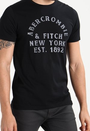 Czarna koszulka z wyblakłym białym napisem "Abercrombie & Fitch New York Est. 1892", noszona przez osobę z tatuażem na lewej ręce.